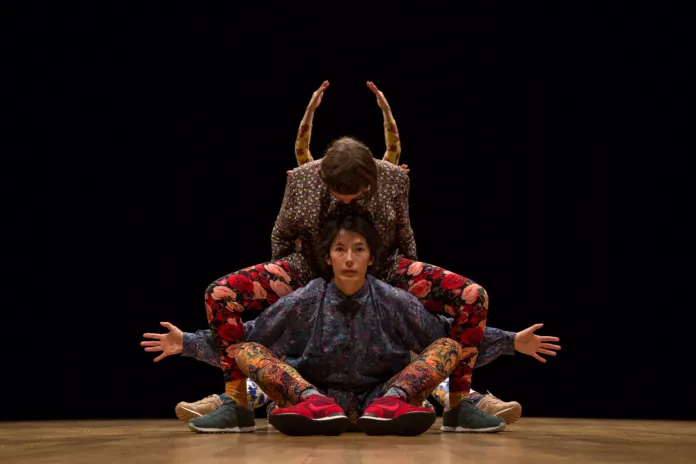Tre danzatori in una composizione simmetrica, con sfondo nero e pavimento in legno.