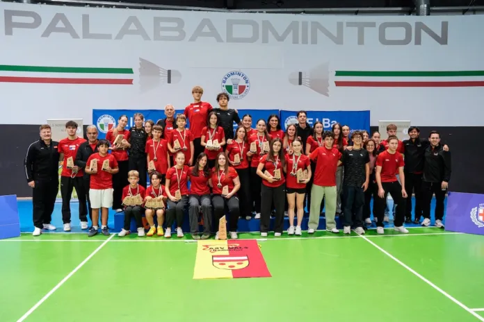Atleti e altre persone posano in gruppo su un campo da badminton, davanti a una rete.