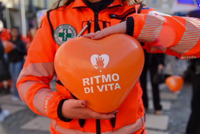 Persone in divisa arancione inginocchiate davanti a manichini per esercitarsi alla rianimazione cardiopolmonare. Una tiene in mano un cuore arancione con la scritta «Ritmo di Vita».