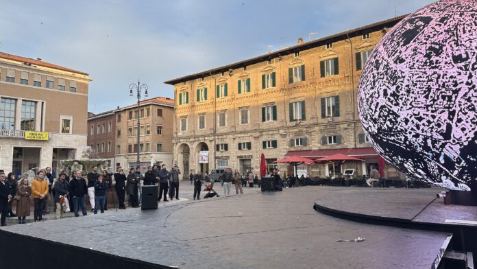 La Biosfera illuminata in una piazza di Pesaro con persone intorno, foto illustrativa dell'opera.