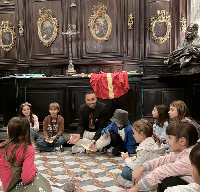 Un gruppo di bambini seduti in cerchio ascolta un adulto in una sala con arredi e decorazioni storiche.