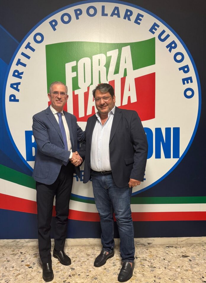 Due uomini stringono la mano davanti a un logo di Forza Italia in un contesto interno di un edificio.