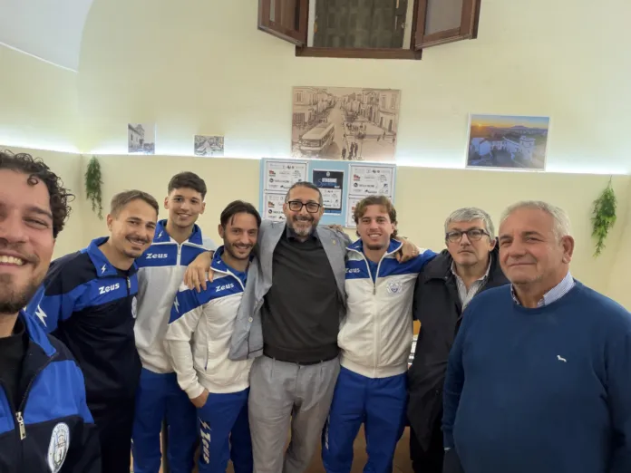 Otto persone posano in una stanza; quattro indossano tute sportive blu e bianche.