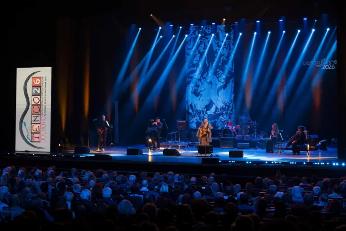 Musicisti si esibiscono su un palco illuminato di blu davanti al pubblico; a sinistra è visibile una scritta Tenco 2026.