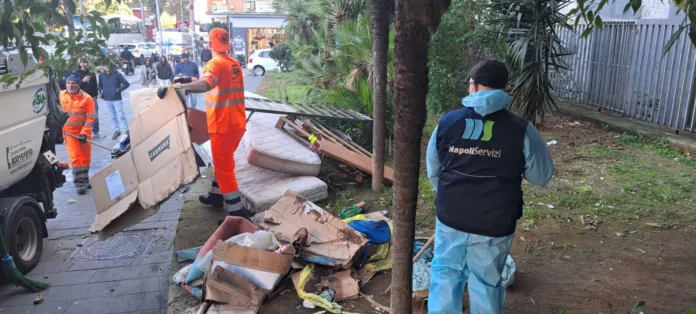 Operatori in abbigliamento da lavoro raccolgono cartoni e altri materiali accanto a un’aiuola; sullo sfondo si vede un veicolo per la raccolta dei rifiuti.