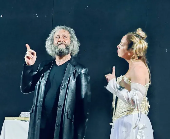 Sul palco, un uomo con giacca scura e una donna in abito chiaro sono rivolti l’uno verso l’altra; entrambi tengono un dito alzato.