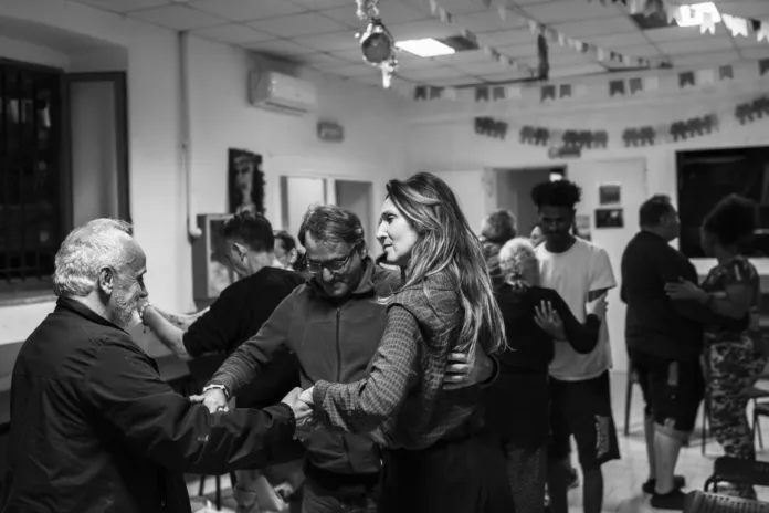 In una stanza, alcune persone interagiscono a coppie; altre sono sullo sfondo.