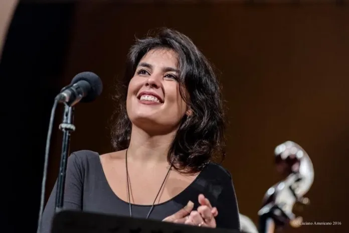 Una donna sul palco, accanto a un microfono; in primo piano si vede parte di uno strumento a fiato.