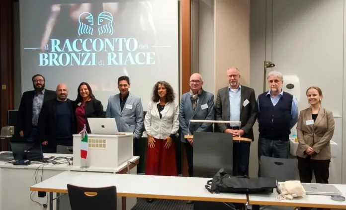 Un gruppo di persone posa in una sala davanti a una proiezione sui Bronzi di Riace; in primo piano si vedono tavoli e attrezzature.