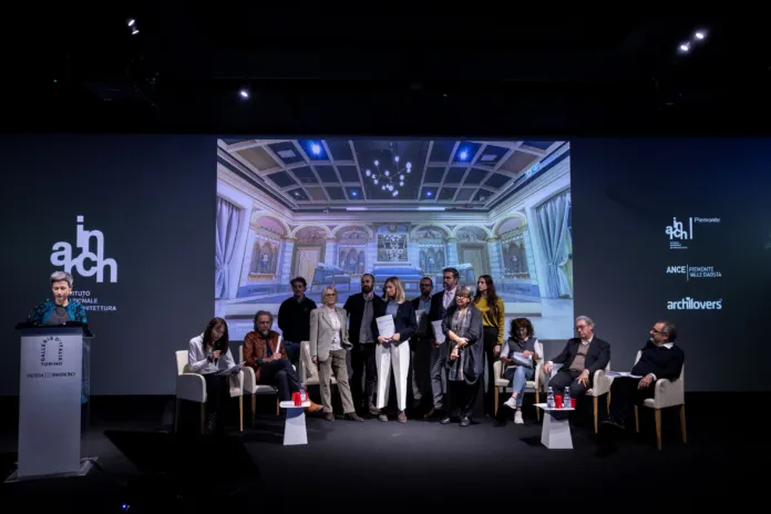 Un gruppo di persone è sul palco; alcune sono in piedi al centro e altre siedono ai lati. Sullo schermo alle loro spalle è proiettata una sala con decorazioni murali.