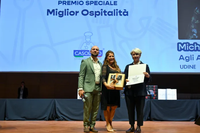 Tre persone posano sul palco; una tiene in mano una targa e un’altra un attestato. Sullo schermo compare la dicitura “Premio speciale Miglior Ospitalità”.