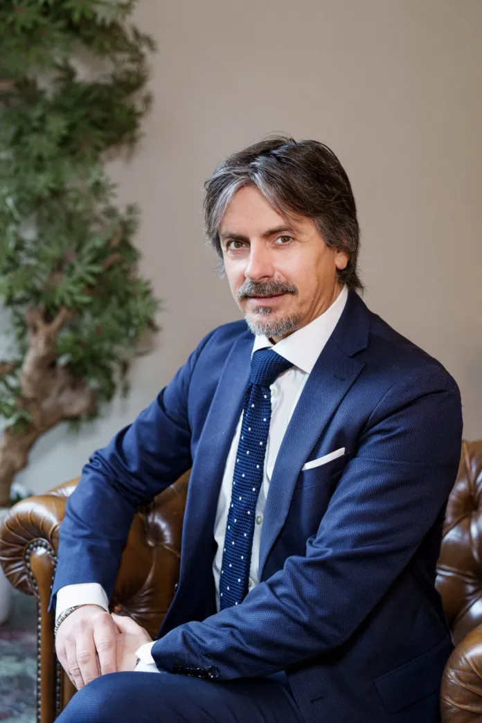 Un uomo con capelli scuri e grigi, in abito blu e cravatta, è seduto su una poltrona in un ambiente interno.