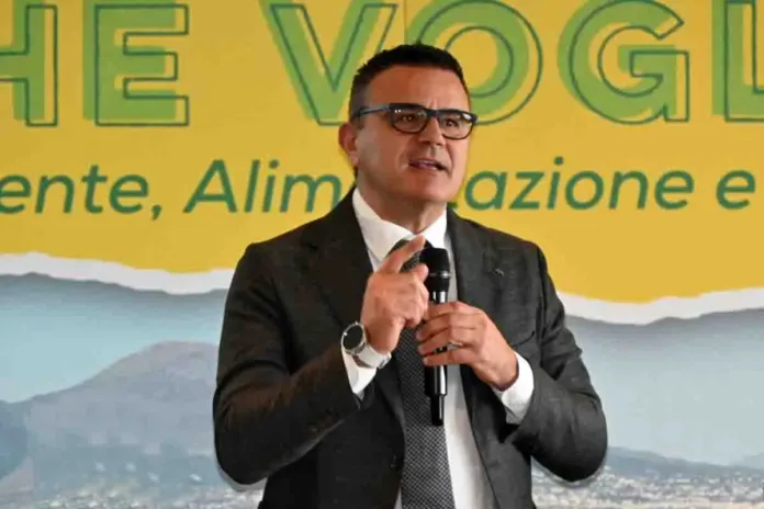 Uomo con occhiali e abito scuro che tiene un microfono davanti a un fondale giallo e verde.