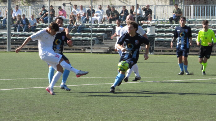 Partita di calcio amatoriale in corso su campo verde con giocatori in azione e arbitro sullo sfondo.