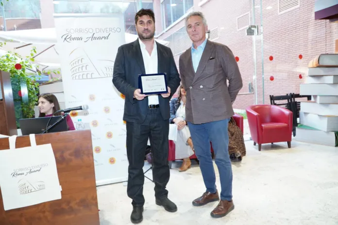 Due uomini in abiti formali, uno dei quali tiene una targa, davanti a un pannello del Sorriso Diverso Roma Award; sullo sfondo si vedono una reception e alcune sedute.