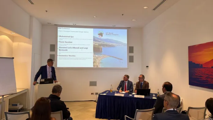 Sala riunioni con un relatore al leggio, una proiezione sullo schermo e partecipanti seduti a un tavolo.