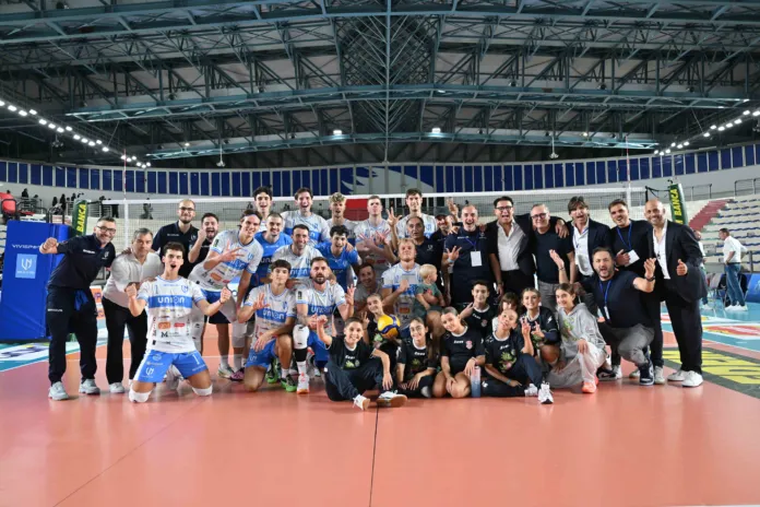 Foto di un gruppo di giocatori e staff della squadra di pallavolo Union Napoli che festeggiano la vittoria, con il pubblico sullo sfondo, in un'ambientazione di un palazzetto dello sport.