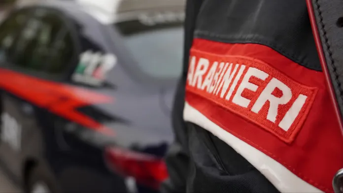 In primo piano una divisa con la scritta «Carabinieri»; sullo sfondo sfocato si vede un veicolo con una striscia rossa.