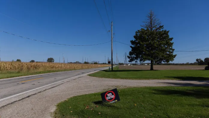Cartello sul prato in una zona rurale, accanto a una strada; sullo sfondo si vedono campi e alberi.