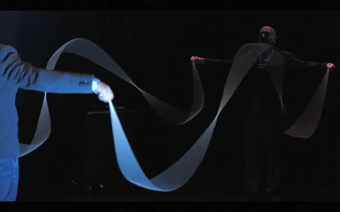 In uno spazio buio, due figure tengono lunghi nastri illuminati di blu.